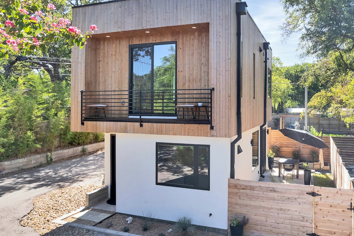 The modern two-story home is showcased with a wooden exterior and large windows. A balcony with two chairs extends from the upper level, offering a view of the surroundings. The entrance is framed by a landscaped area, with a mixture of gravel and plants at the base.