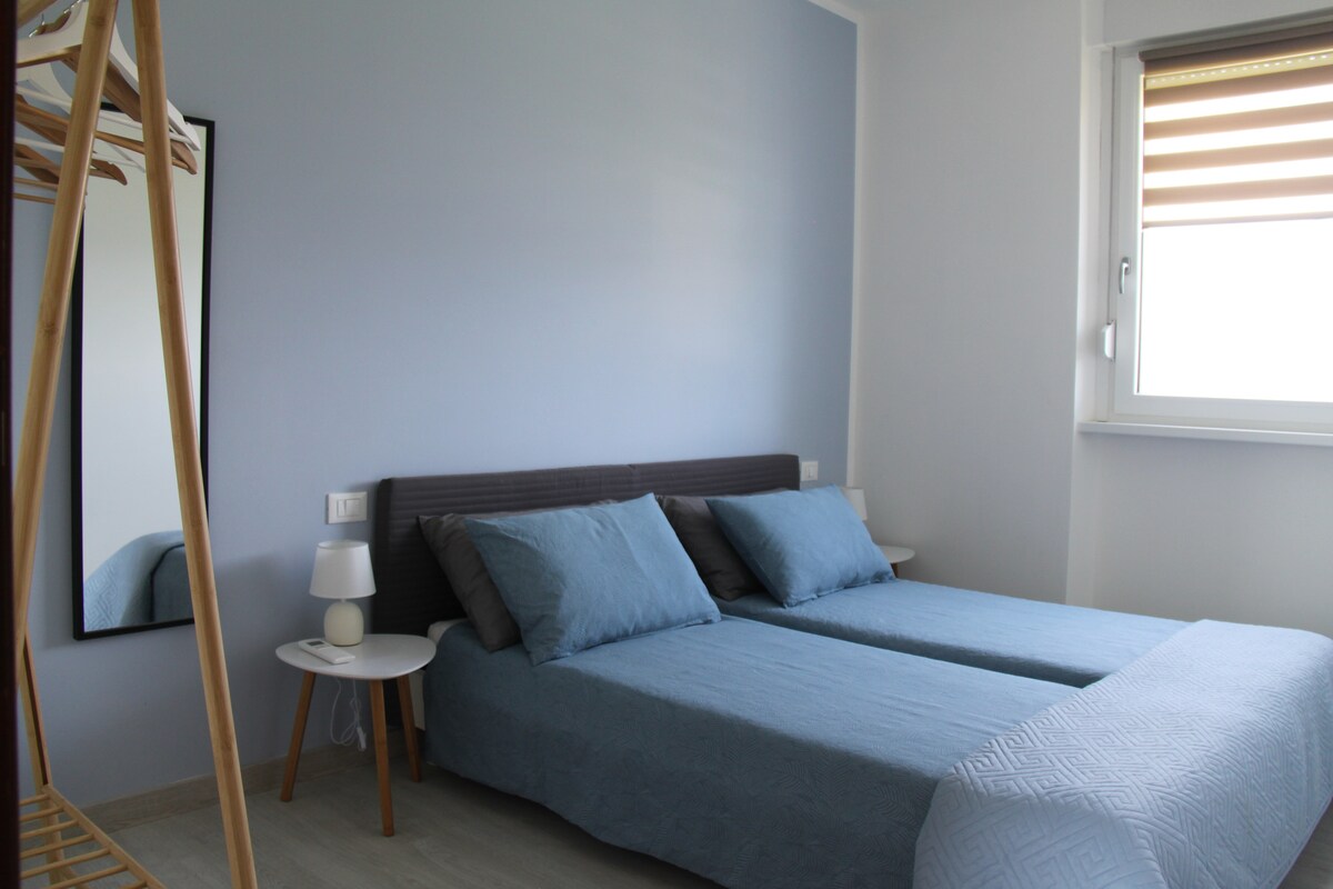 A light and airy bedroom features two single beds dressed in soft blue linens. A bedside table accompanies a contemporary lamp. A tall mirror reflects the space, while a window with dual-tone blinds allows natural light to fill the room.
