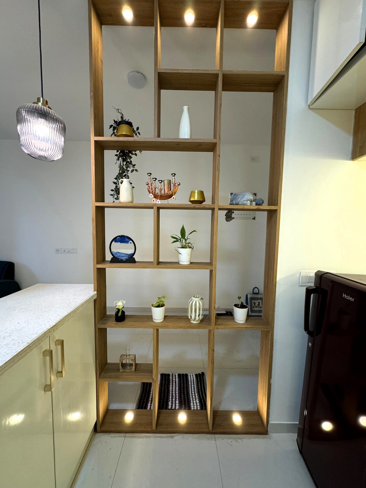 A modern wooden shelving unit is illuminated with soft lights, displaying various decorative items such as vases and plants. The unit is situated near a kitchen area, enhancing the overall aesthetic with its organized arrangement.