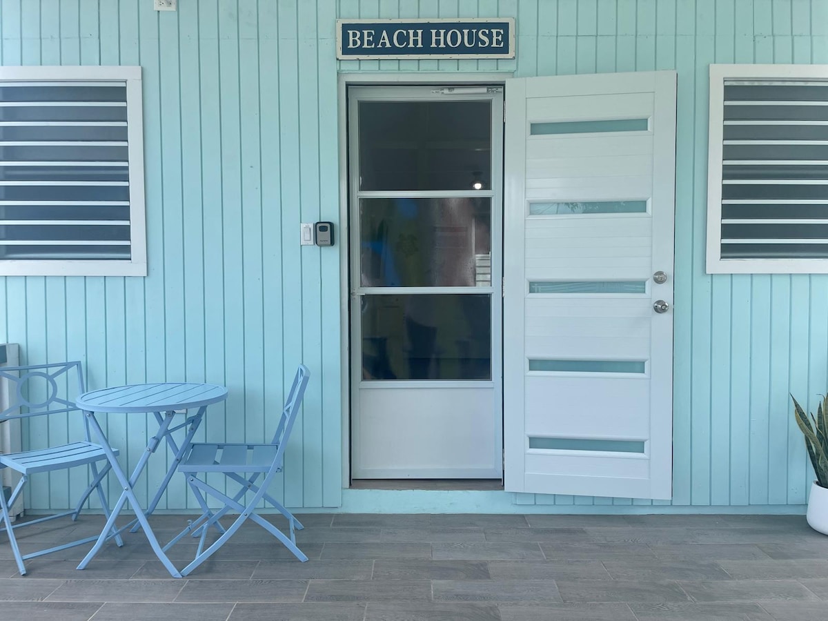 A light blue exterior features a welcoming entrance to the beach house. The door is framed by two windows and a small, round table with two chairs sits nearby, providing a space for relaxation. A potted plant adds a touch of greenery.