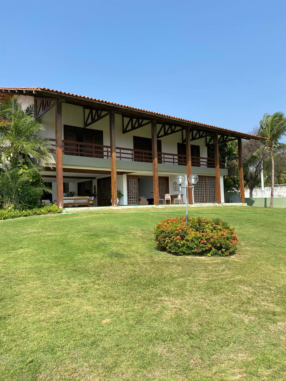 The two-story house features a spacious balcony that overlooks a well-maintained lawn. Sunlight illuminates the structure, accentuating the wooden beams and large windows. Lush greenery and colorful flowering plants are present in the foreground, contributing to a serene outdoor environment.