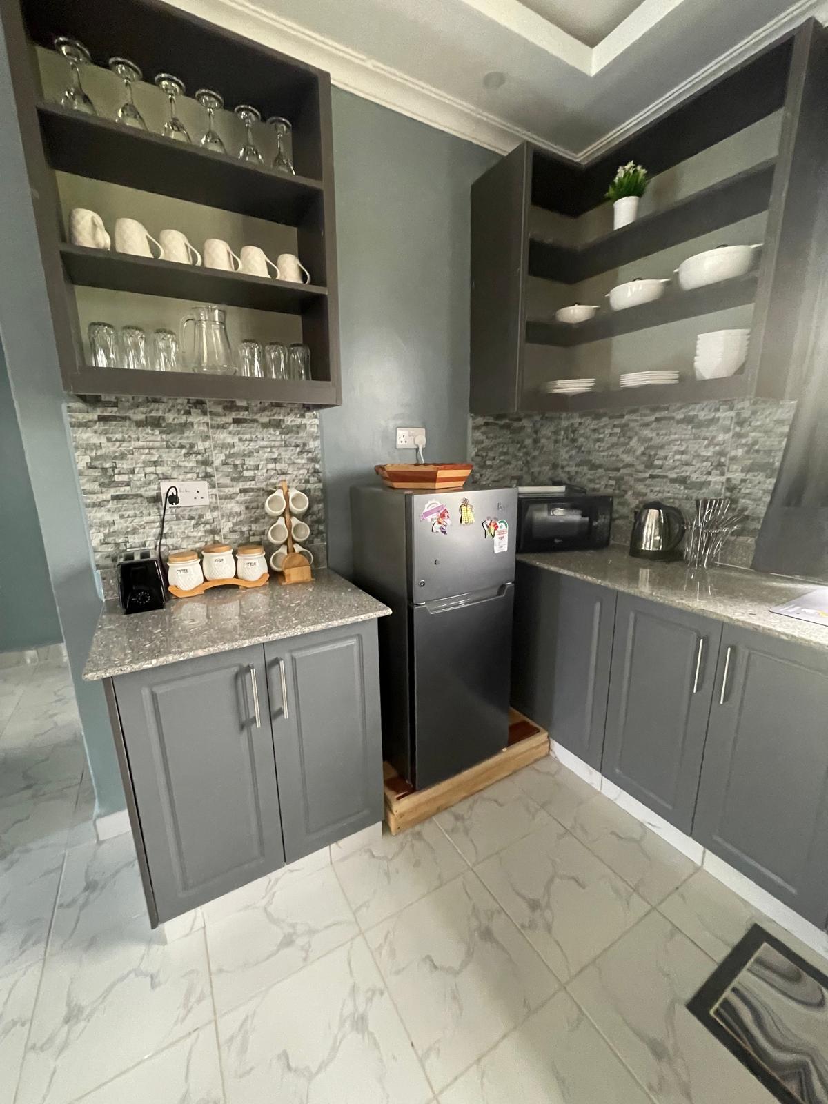 A functional kitchen showcases modern cabinetry with ample storage, glassware, and dishware neatly arranged on open shelves. A refrigerator, microwave, and kettle are present, situated against a textured stone backsplash. Countertop space is available for meal preparation, with a tiled floor enhancing the space's overall design.