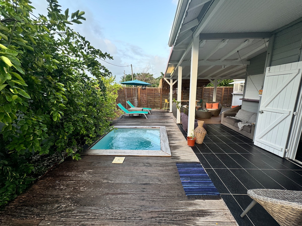 An outdoor area features a small spa surrounded by wooden decking. Sun loungers in vibrant colors are positioned nearby, while a shaded seating area is visible in the background. Lush greenery enhances the sense of privacy in this tranquil space.