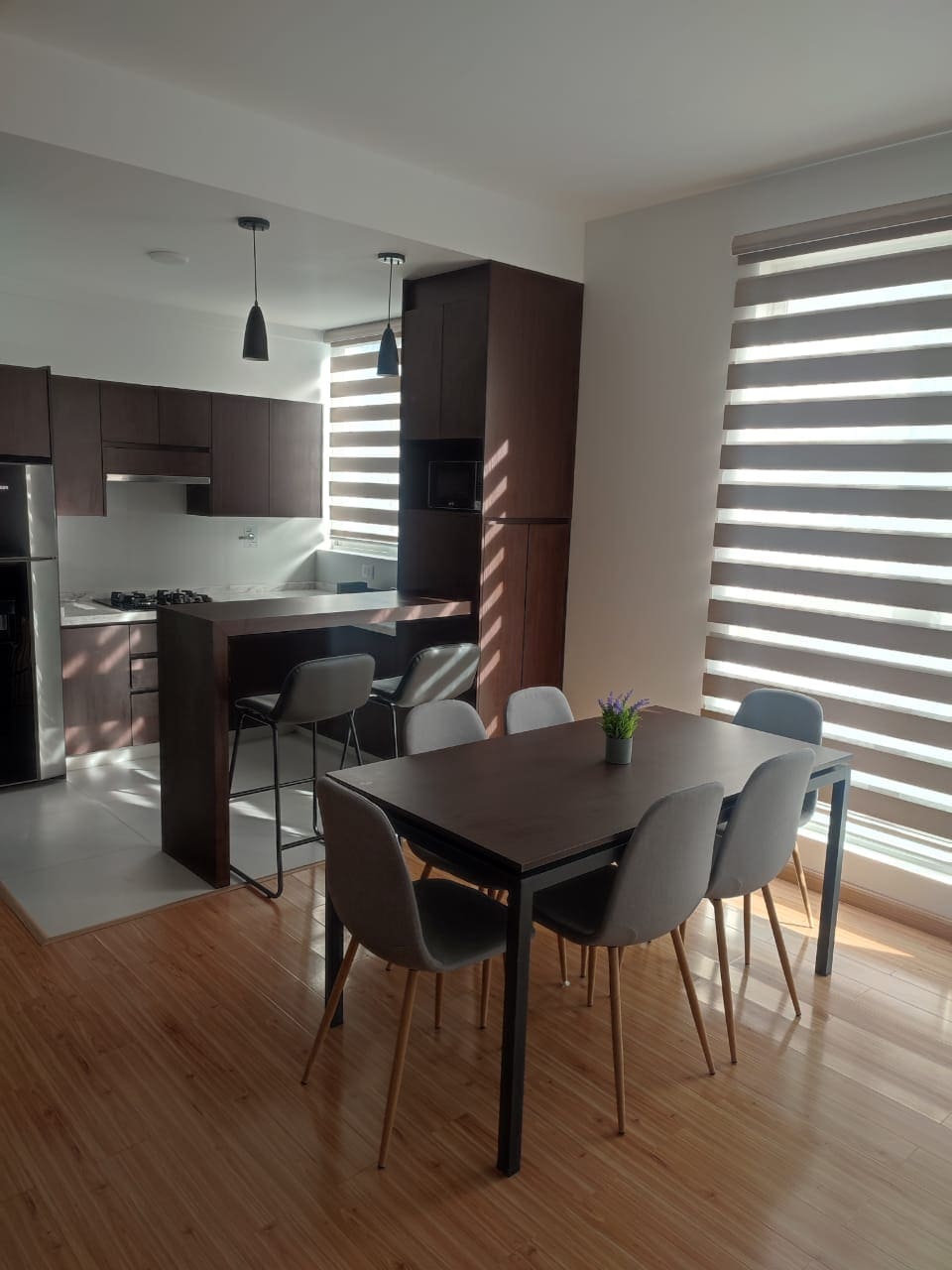 An open dining area features a dark wooden table surrounded by six grey chairs. A modern kitchen is visible in the background, equipped with dark cabinetry and stainless steel appliances. Light from the large window creates a bright atmosphere, partially obscured by striped blinds.