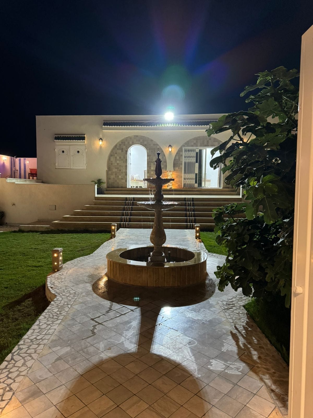 A tranquil courtyard features a central fountain with water flowing gently. Surrounding the fountain, stone pathways lead to steps ascending towards a well-lit entrance. Lush greenery adds to the serene ambiance, complemented by soft lighting that enhances the evening atmosphere.