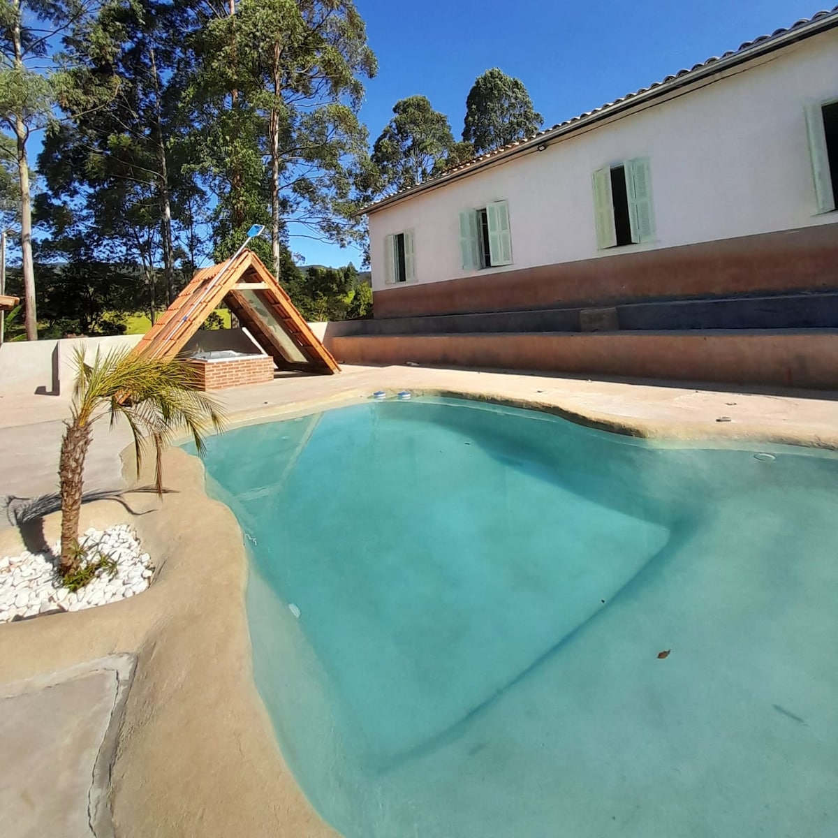 A serene pool is shown surrounded by a smooth deck. A wooden play structure with a sloped roof is visible nearby. Sunlight reflects off the clear water, with green trees in the background and a white building featuring green shutters.