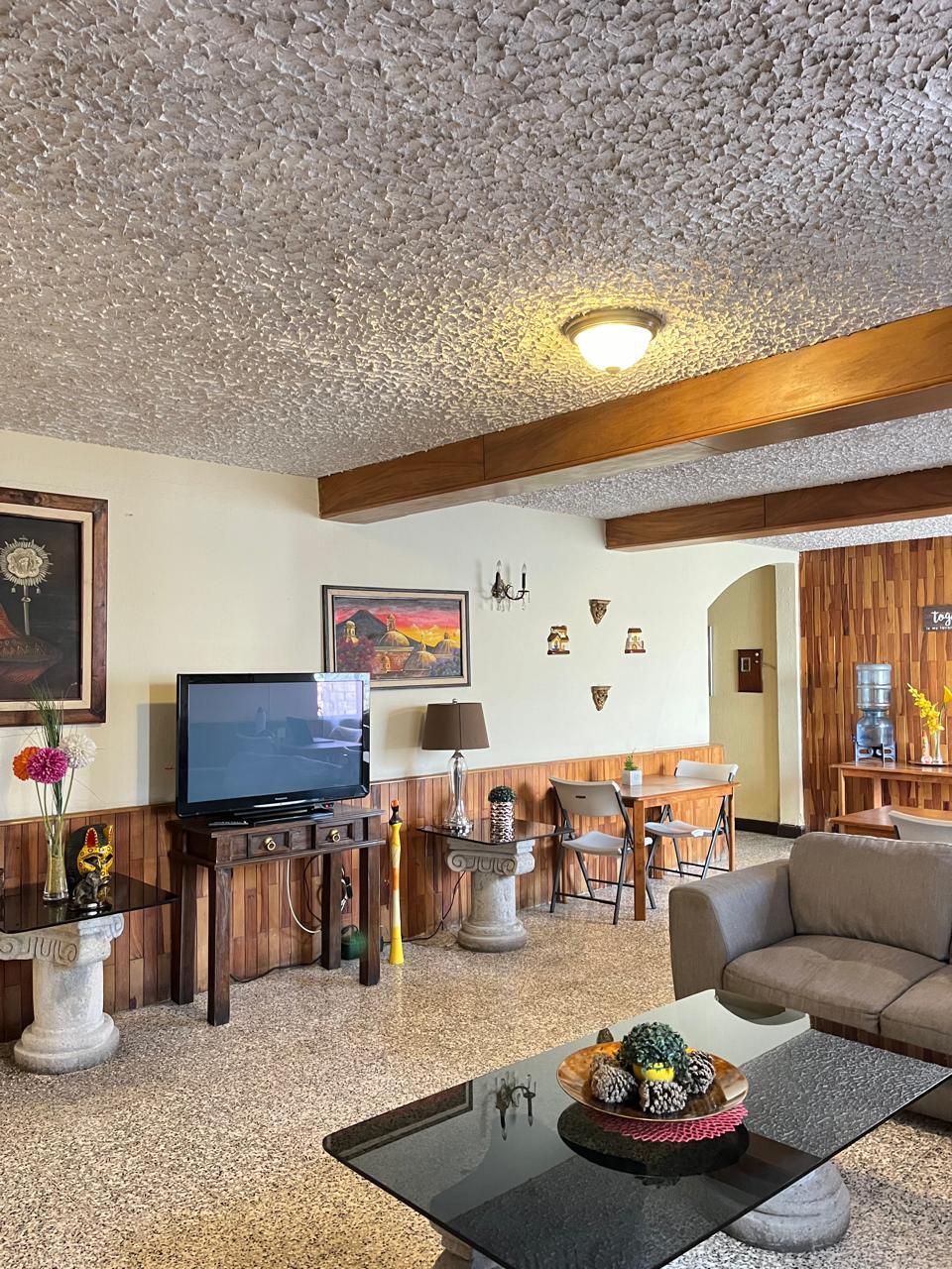 An open living area is showcased with a comfortable sofa and a black glass coffee table, complemented by decorative fruit. A television is positioned against one wall, with wooden accents and artwork providing a homey touch. A dining area is glimpsed in the background.