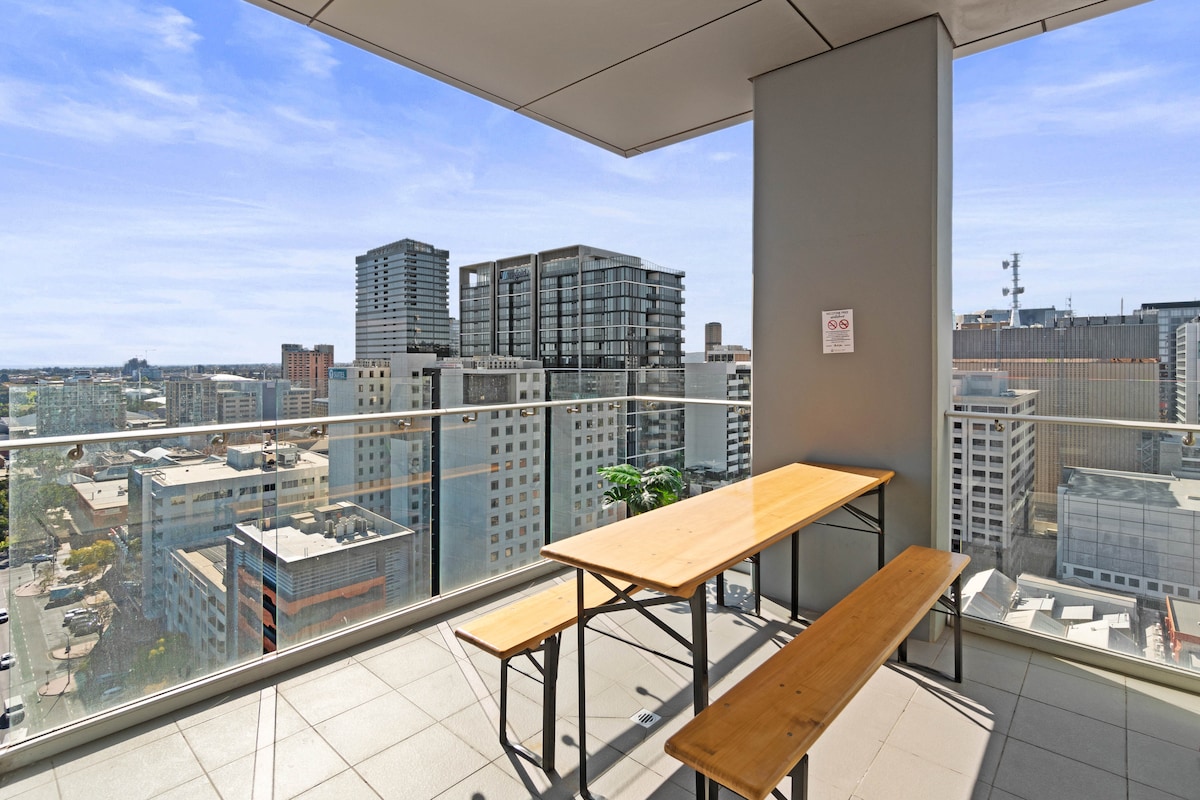 A private balcony features a wooden table and bench seating with views overlooking the city skyline and Light Square. The expansive glass railing provides unobstructed panoramic sights of the surrounding urban landscape.