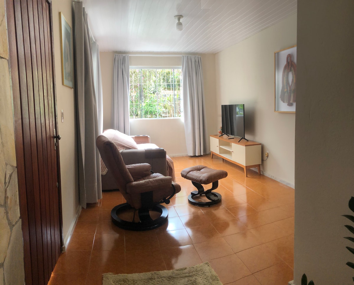 A living area is shown with a light, neutral color scheme. A comfortable seating arrangement includes a recliner and an ottoman facing a TV unit. Natural light filters through a large window adorned with sheer curtains, illuminating the glossy tiled floor.