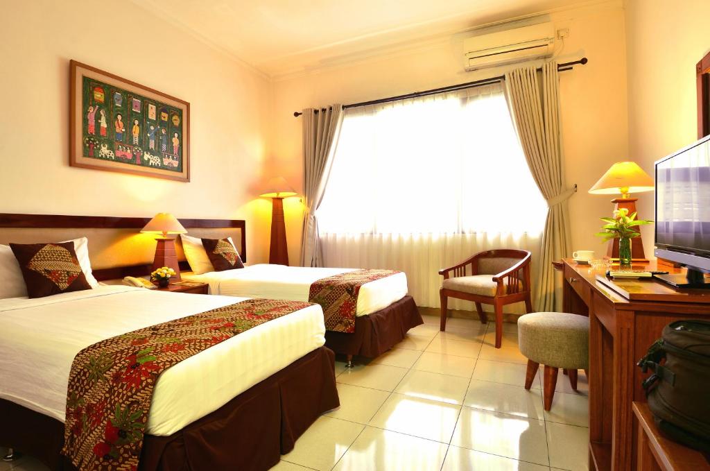 The room features two single beds with patterned bedspreads, complemented by wooden furniture. A window lined with light curtains allows natural light to brighten the space. A flat-screen TV, a desk, and a chair are present, along with bedside lamps providing additional illumination.