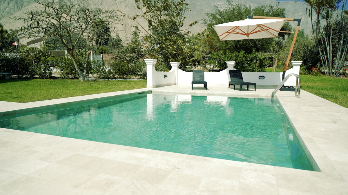 A serene outdoor space features a clear swimming pool surrounded by light-colored stone tiles. A shaded seating area with two dark chairs and a sun umbrella is visible beside the pool, while lush greenery creates a tranquil backdrop.