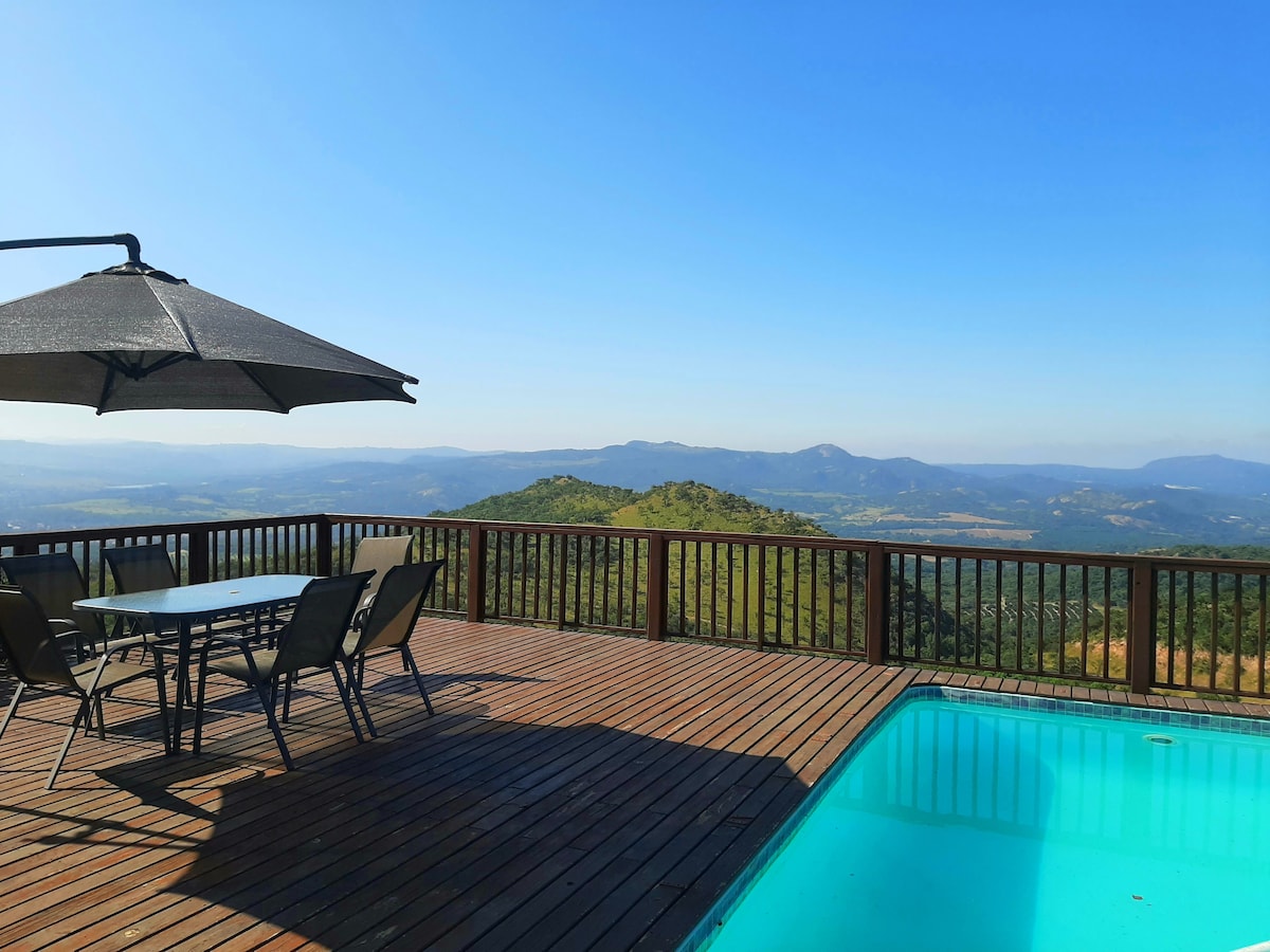A wooden deck features a swimming pool and a dining table with chairs, bordered by a panoramic view of rolling hills and valleys under a clear blue sky. A large umbrella provides shade over the seating area.
