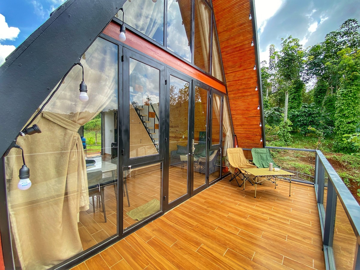 A spacious balcony features large glass doors that provide an unobstructed view of the surrounding greenery. The area is furnished with outdoor seating, including a folding chair and a lounge chair, inviting relaxation while enjoying the natural surroundings.