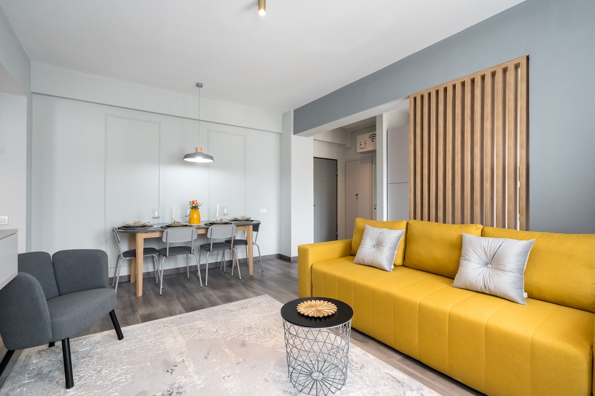 A modern living area is presented with a bright yellow sofa, complemented by a patterned area rug and a stylish coffee table. A dining table for four is visible in the background, softly illuminated by an overhead light fixture.