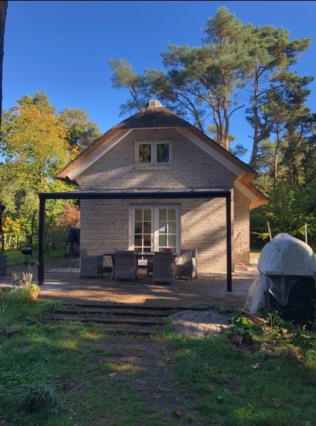 A modern villa is set in a forest area, featuring a thatched roof and large windows. A covered outdoor seating area is located in front, furnished with a dining table and chairs. Surrounding greenery enhances the nature-inspired setting.