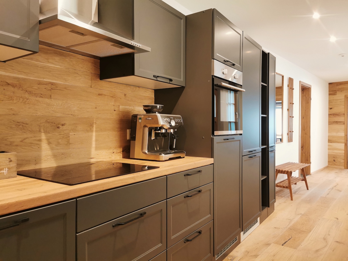A modern kitchen is presented with sleek black cabinetry and wooden accents. An espresso machine is prominently displayed on the countertop, along with a stovetop and built-in oven. Light-colored wooden flooring adds warmth to the space, while natural light brightens the area.