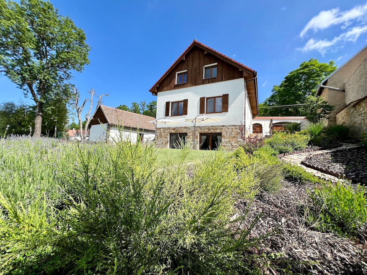 A charming countryside chalupa is showcased against a bright blue sky, framed by lush greenery and blooming lavender. The building features a wooden upper floor with windows and a stone lower level, surrounded by a manicured lawn and well-kept shrubs.