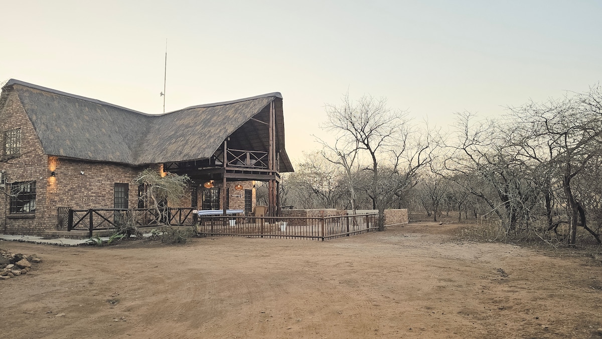 A rustic brick lodge is set against a backdrop of sparse trees and dry earth. The structure features a distinctive thatched roof and a welcoming porch with a railing. The surrounding area exhibits a secluded and tranquil landscape, showcasing nature's simplicity.