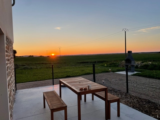 An outdoor terrace features a wooden table with benches, set against a backdrop of vibrant sunset colors. The open view includes grassy fields, while a barbecue area is visible in the distance. The space is enclosed by a fence, ensuring privacy.