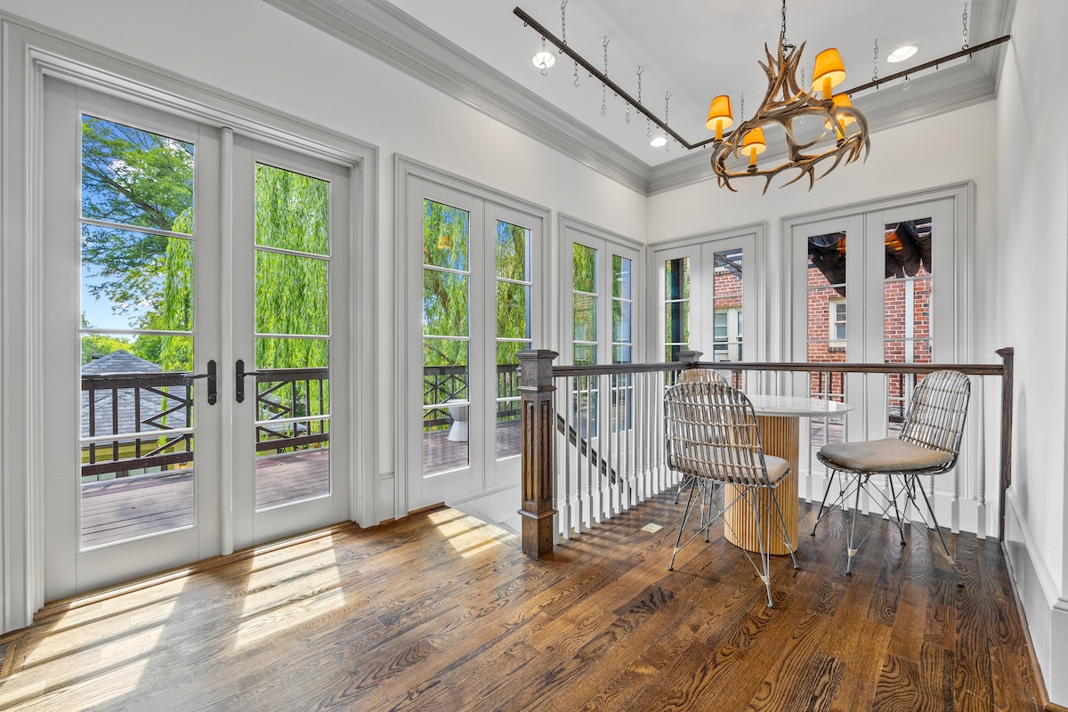 A sunlit balcony space features large glass doors offering a view of the surroundings. Two stylish chairs and a small table are positioned on the polished wooden floor, which is complemented by natural light streaming in from multiple windows.