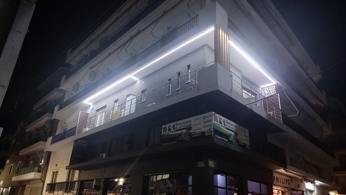 An illuminated three-story building is seen at night, featuring a modern facade with balcony railings. Soft white light highlights the architecture, while shops occupy the ground level. The surrounding area is urban, with street elements visible.