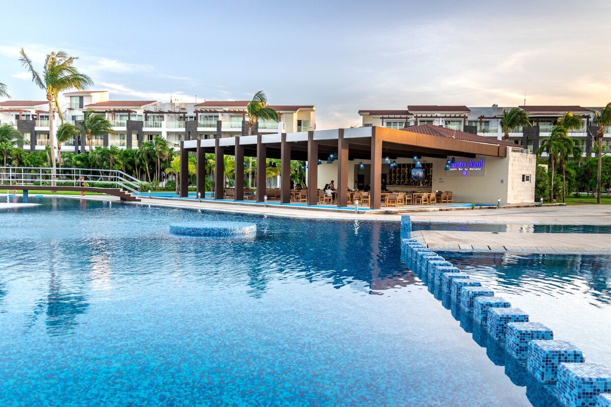 A large swimming pool reflects the sky, with a modern poolside bar and dining area visible in the background. Lush green landscaping surrounds the space, enhancing the inviting atmosphere for relaxation and enjoyment.
