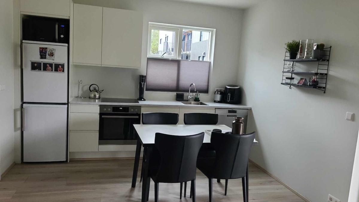 A modern kitchen features sleek cabinetry and stainless steel appliances, including a refrigerator and an oven. A dining table is set with four black chairs, and a window provides natural light. Open shelving displays a few decorative items, enhancing the functional space.