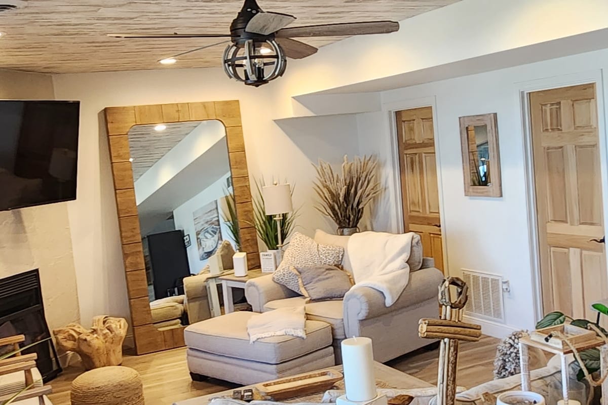 A welcoming living area is shown, featuring a light-colored sofa and a stylish ottoman. A large mirror reflects the space, while decorative plants and candles are arranged on a natural wood table. Light wooden flooring complements the neutral decor, creating an inviting atmosphere.