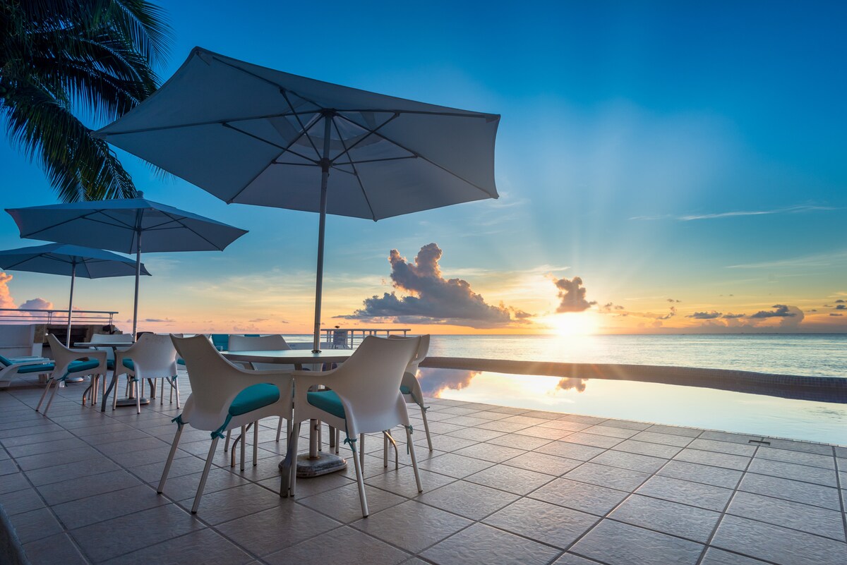 A tranquil outdoor terrace is depicted, featuring several white chairs and tables under large umbrellas. The infinity pool reflects the vibrant colors of the sunset, while the ocean stretches into the distance, creating an expansive view of the horizon.
