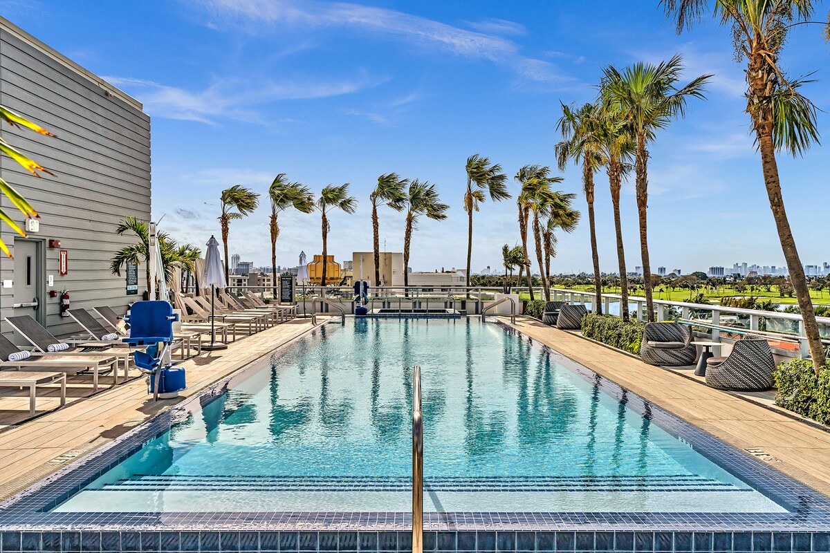 A spacious outdoor pool area is surrounded by lounge chairs and palm trees swaying gently in the breeze. The clear blue water reflects the sky, creating a serene atmosphere. The deck offers ample space for relaxation, with views extending towards the skyline.