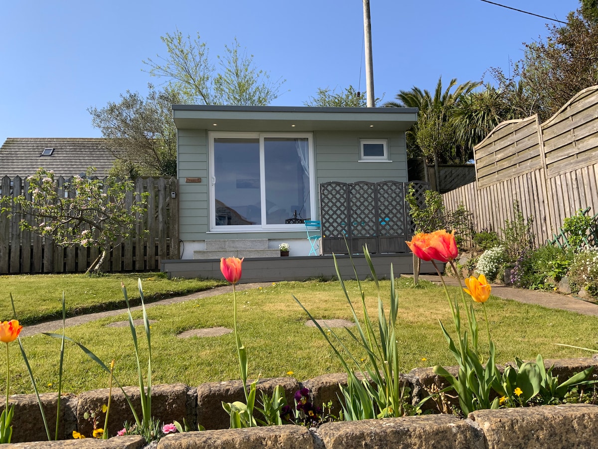 A charming garden hut is framed by colorful tulips and greenery. Large glass doors allow natural light to fill the space, and a small decorative table is positioned to the side. The overall setting offers a serene outdoor view with a manicured lawn.