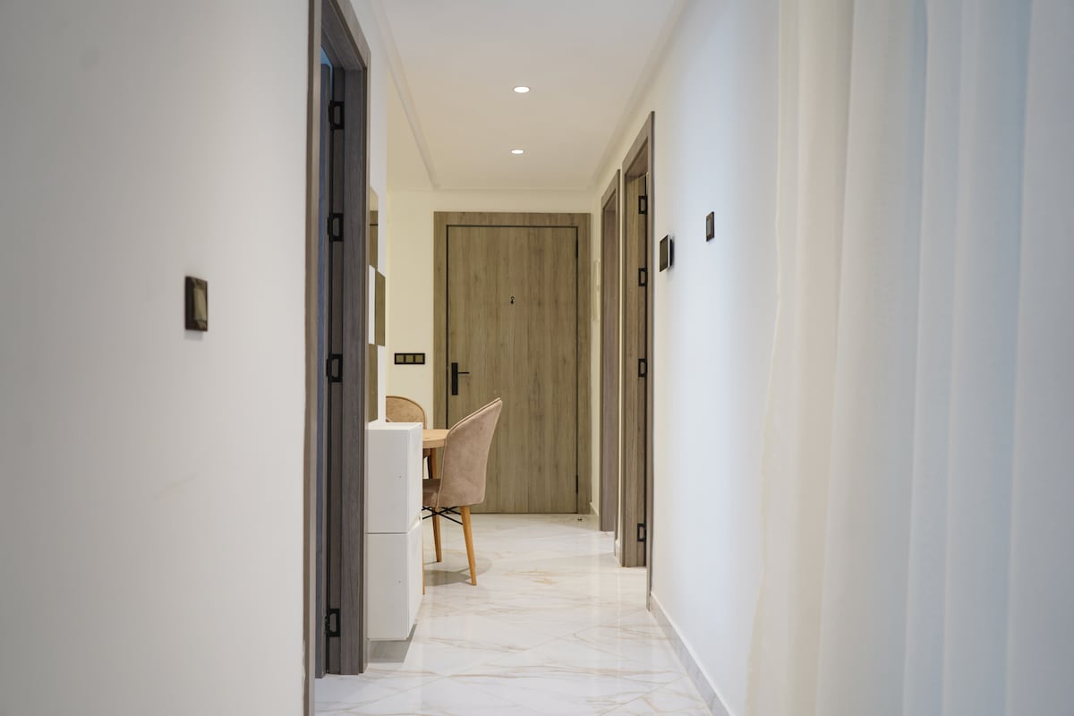 A bright hallway is visible, featuring sleek marble flooring and well-lit walls. Multiple doors lead to various rooms, while a small desk with a chair is positioned against one wall, creating a functional and inviting space.