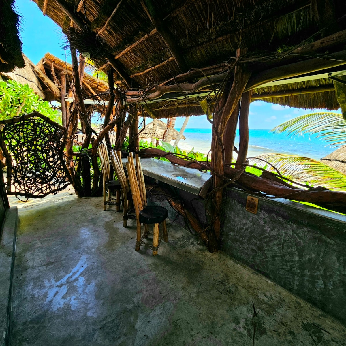 An open terrace features a rustic wooden dining table surrounded by tall chairs. Natural materials blend seamlessly with the ocean view, which can be seen through large openings. Nearby, a cozy hammock is positioned for relaxation, enhancing the tranquil setting.