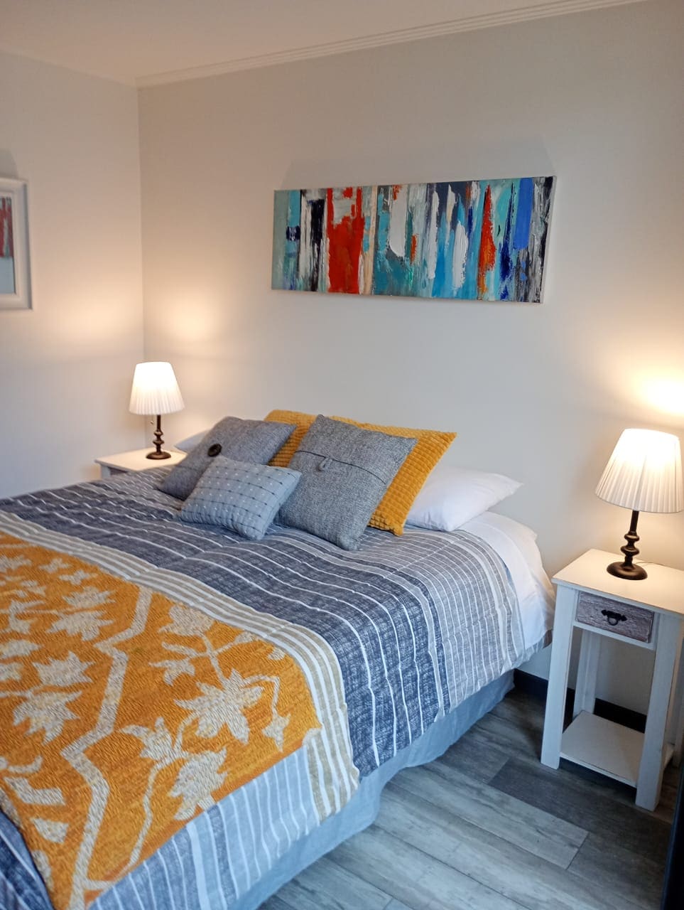 A neatly made bed is positioned against a neutral-colored wall, featuring a striped blanket and decorative pillows. Two bedside lamps provide soft lighting, and a colorful abstract art piece hangs above the headboard, enhancing the room's calm atmosphere.