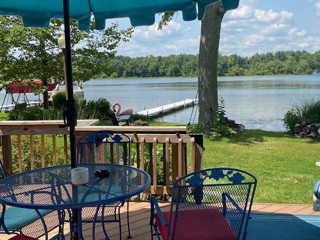 A deck area is shown with a round table and four colorful metal chairs. A green umbrella provides shade, and the view reveals a tranquil lake with a wooden dock extending into the water. Lush greenery surrounds the waterfront.