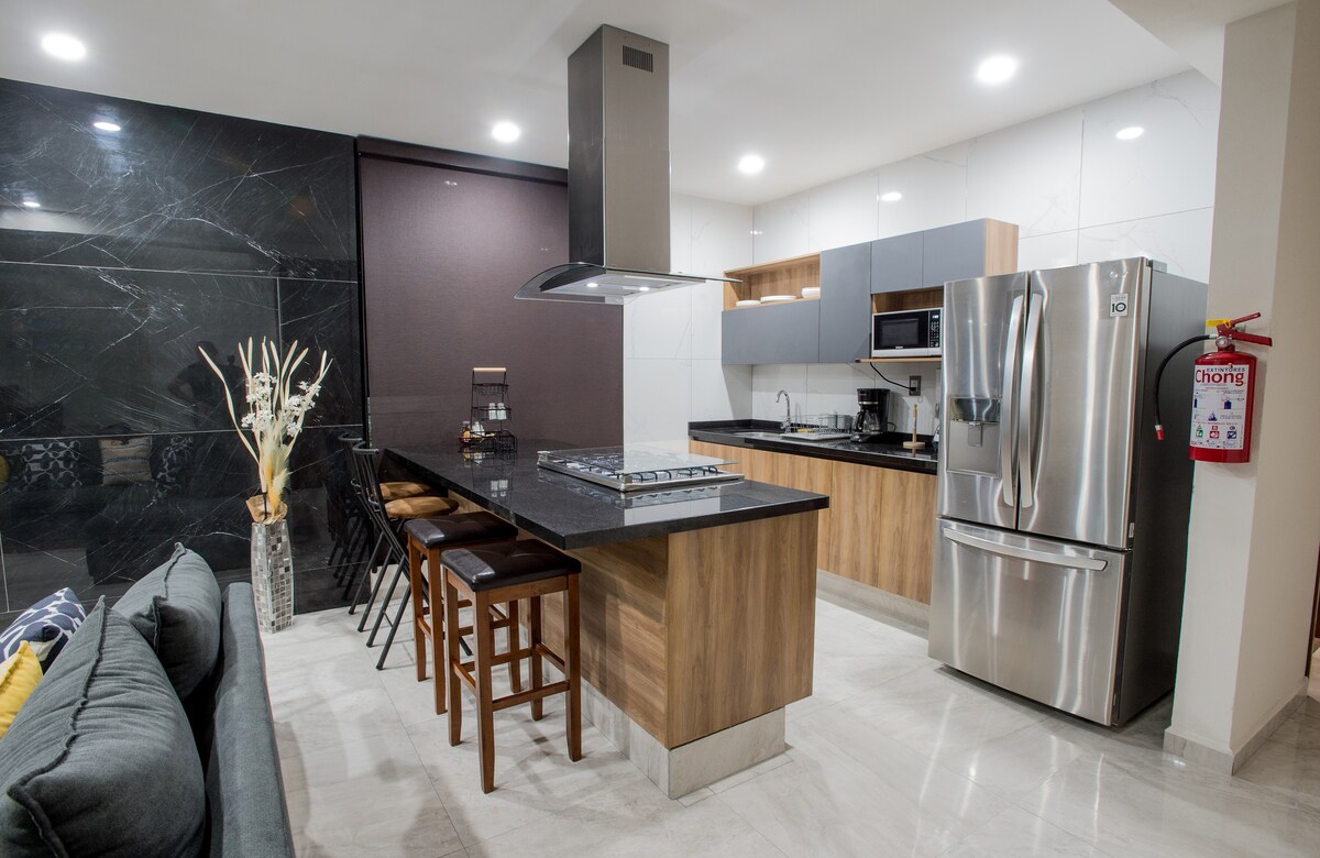 A modern kitchen is highlighted, featuring sleek cabinetry and a large island with seating for four. Stainless steel appliances are positioned alongside a black countertop, while decorative elements include a vase with dried flowers. Soft lighting enhances the clean, contemporary design.