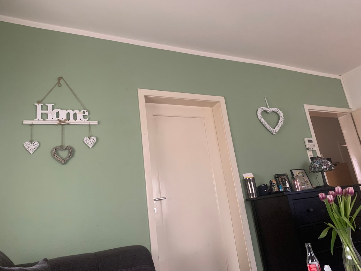 A green wall features decorative elements, including a wooden 'Home' sign and two heart-shaped wall hangings. A closed white door leads to another space, accompanied by a dark-colored cabinet displaying photographs and decorative items.