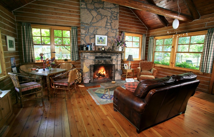 A warm interior features a stone fireplace as the focal point, surrounded by comfortable seating and a dining area with wooden furniture. Large windows allow natural light to illuminate the space, enhancing the rustic charm of the wooden walls and ceiling.
