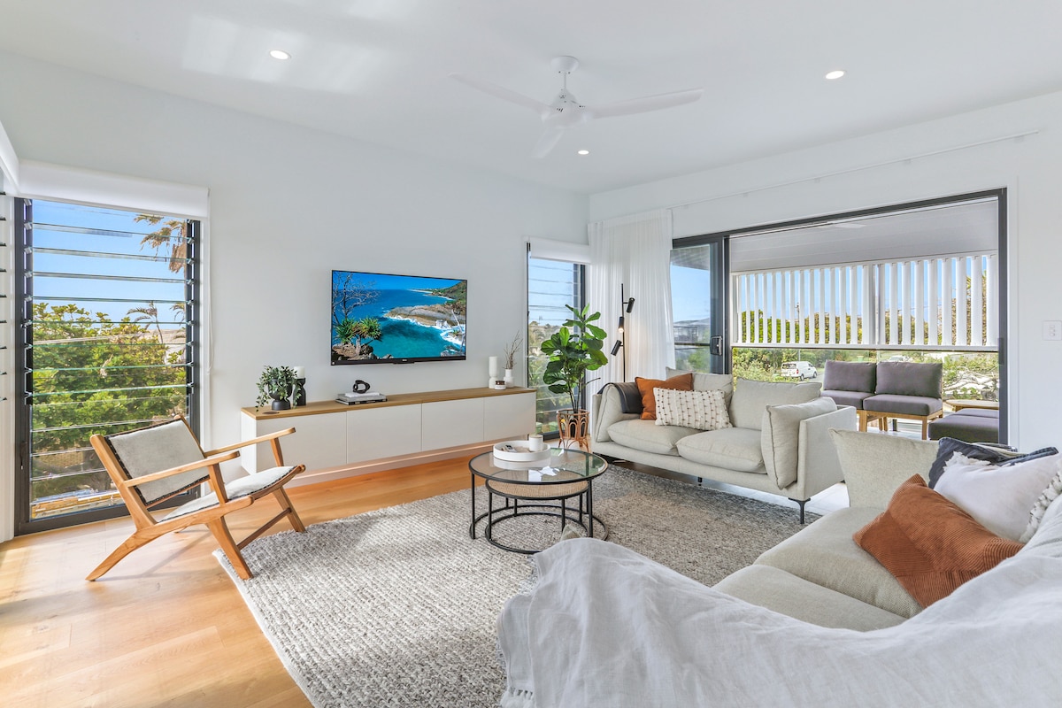 A bright living area showcases large sliding glass doors leading to the outdoor space. The room features a cozy sectional sofa with decorative cushions, an accent chair, and a round coffee table atop a textured rug. A wall-mounted TV and a decorative plant enhance the modern aesthetic.