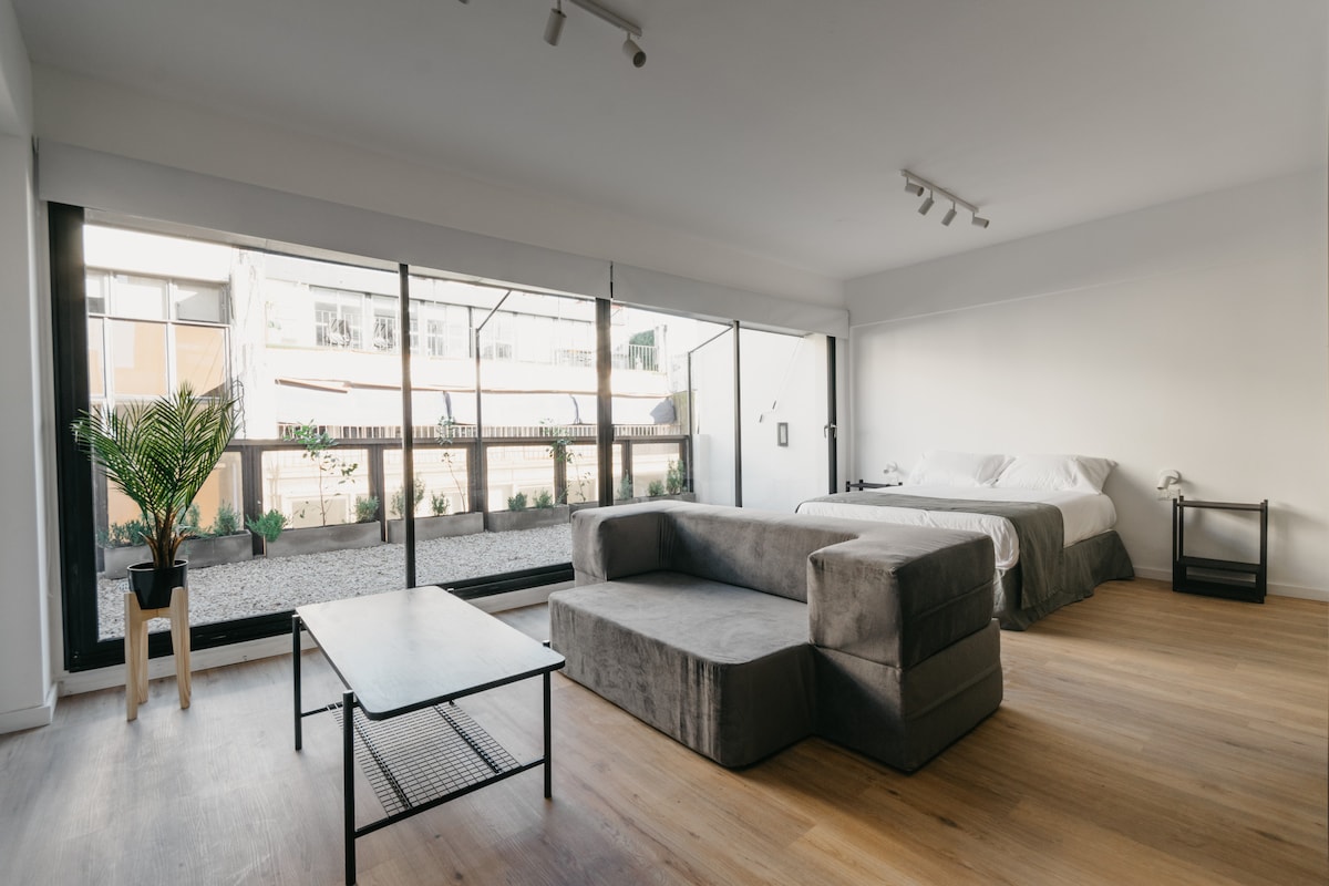 A spacious room features a comfortable bed and a large sectional sofa. A glass door provides access to an outdoor area, while natural light fills the room. Simple furnishings include a small black coffee table and a potted plant, adding a touch of greenery.