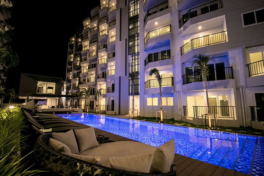 The outdoor area features a long, illuminated swimming pool with a tranquil blue hue, bordered by stylish loungers. The modern condominium buildings rise in the background, with numerous bright windows reflecting the night sky.