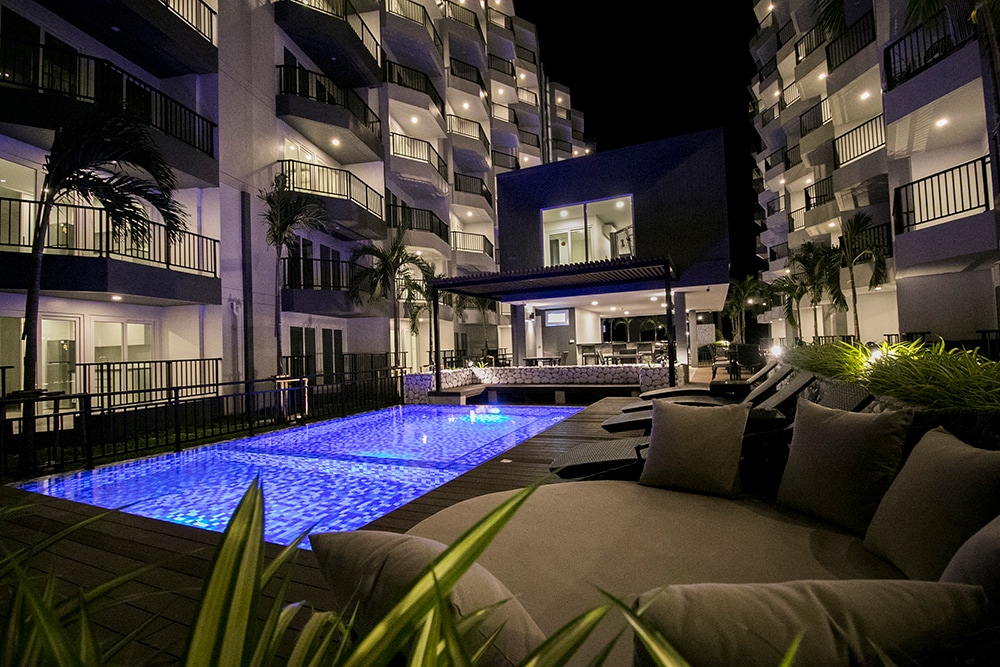 The illuminated swimming pool reflects the night sky, surrounded by comfortable lounge chairs and palm trees. The modern condominium building stands in the background, with spacious balconies visible on each floor, creating a relaxed and inviting outdoor environment.