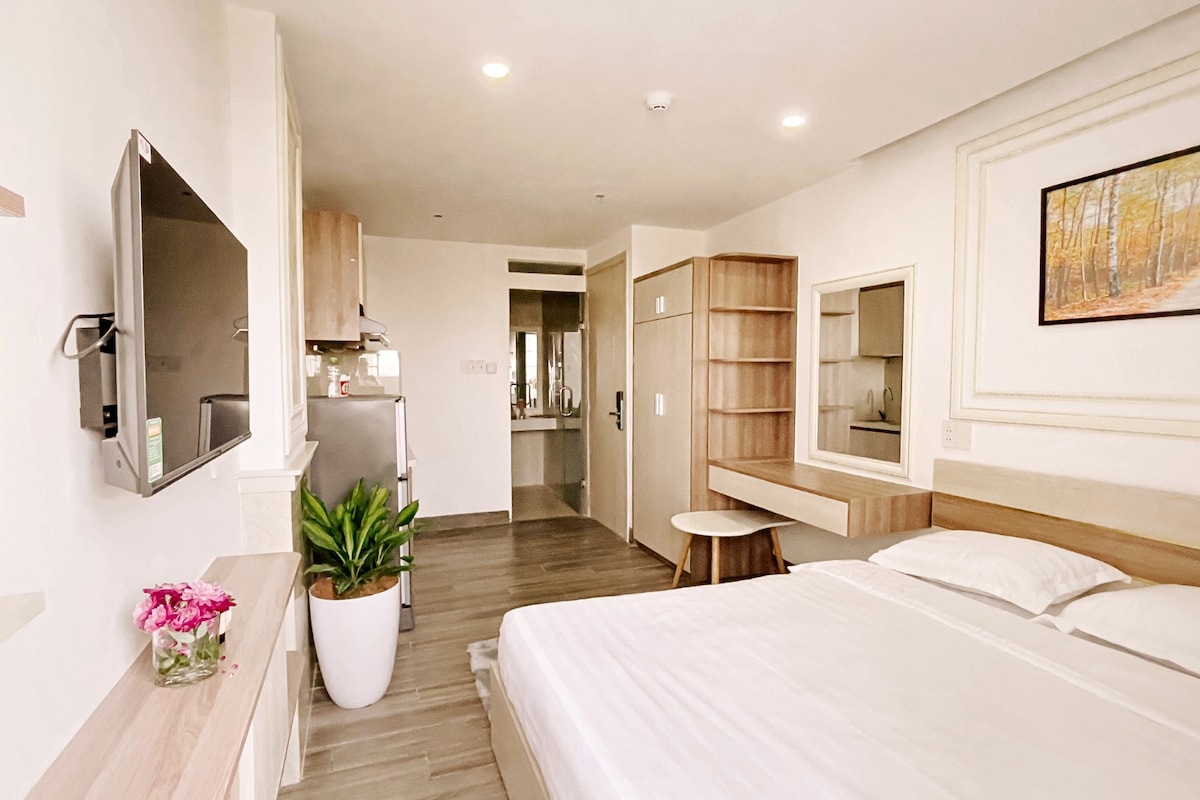 A minimalist bedroom features a spacious bed with white linens. A modern television is mounted on the wall opposite, while a small dining table and a potted plant add charm. An open kitchenette is visible in the corner, with a mirror reflecting the layout.