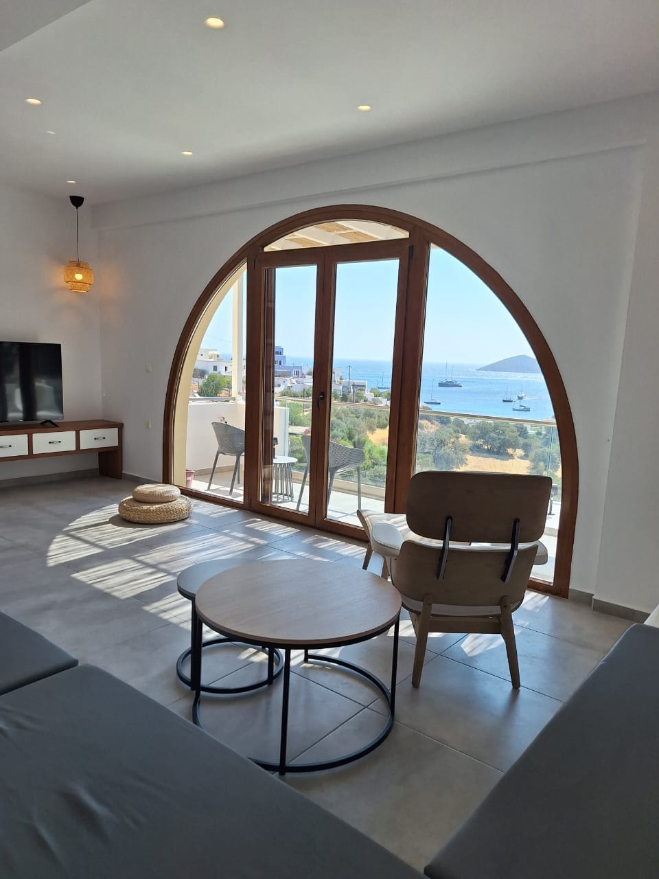 A large arched window frames a scenic view of the bay, bringing natural light into the living area. A comfortable chair and small round table are positioned nearby, adding to the relaxed atmosphere. The space features neutral tones and modern furnishings.