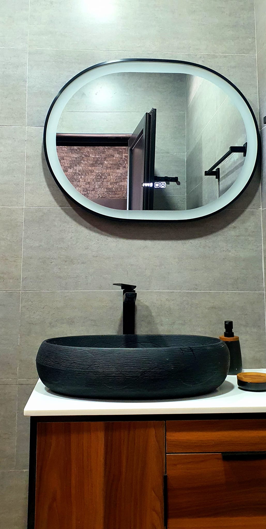 A modern bathroom features a circular mirror above a stylish, dark bowl sink. A sleek black faucet complements the minimalist design. The countertop is made of light-colored material, while the cabinetry below is finished in warm tones. A partial view of a shower area is visible in the background.