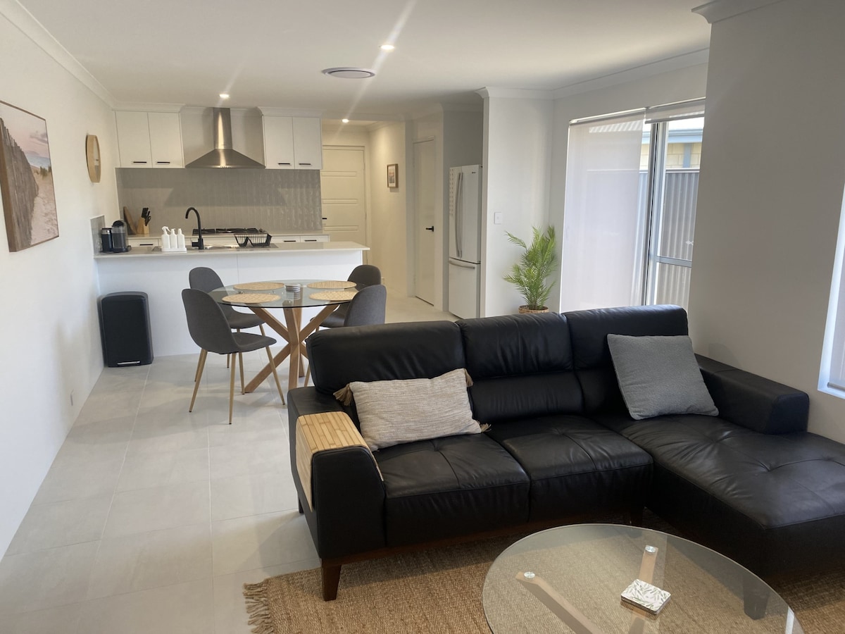 A modern open-plan living and dining area showcases a comfortable black sectional sofa and a glass coffee table. Towards the background, a sleek kitchen is outlined and a round dining table with four chairs is visible. Natural light filters through the windows, enhancing the spacious atmosphere.
