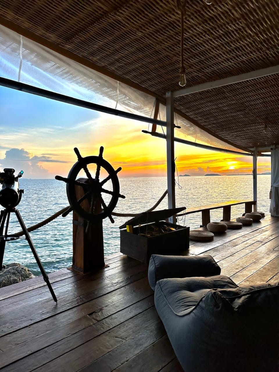 An inviting outdoor space features a wooden steering wheel capturing the essence of adventure, overlooking the ocean. The setting sun casts warm hues across the tranquil water. A cozy seating area with plush cushions is arranged beside a table, enhancing the relaxed atmosphere.