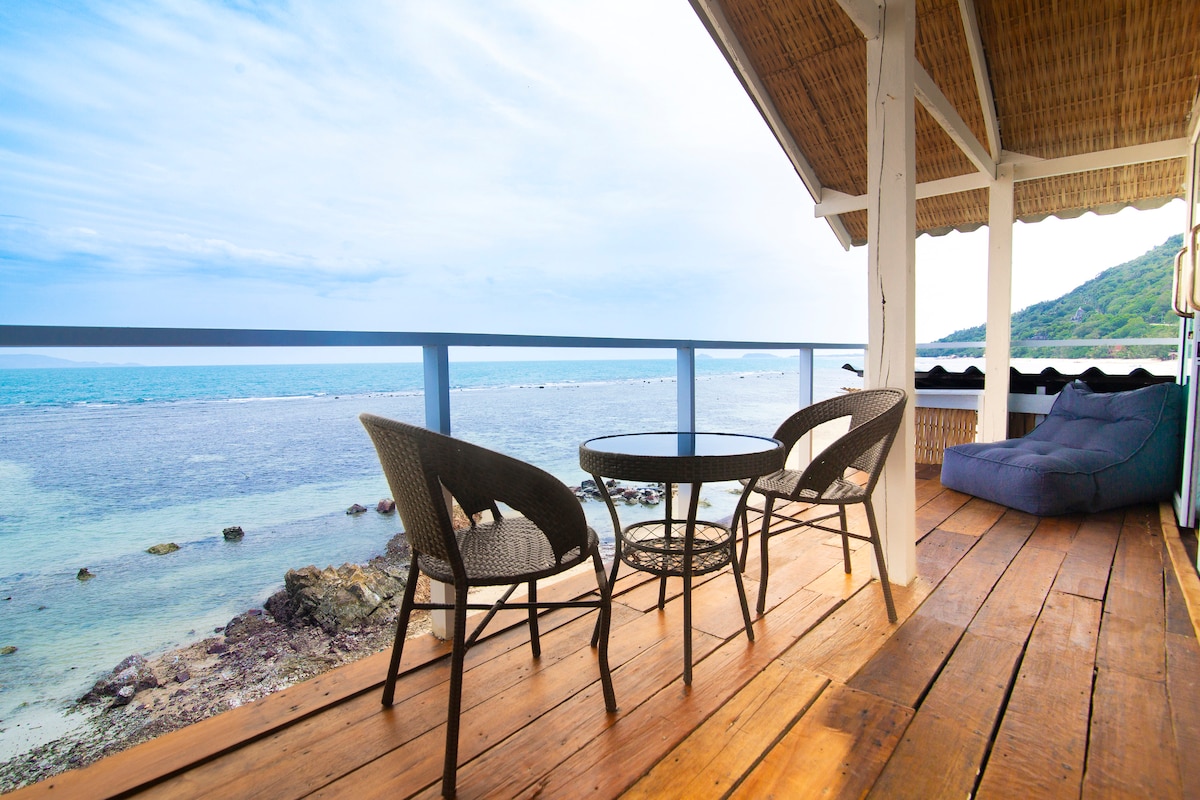 An outdoor terrace showcases a scenic view of the ocean with gentle waves. A round table and four cozy chairs are positioned on the wooden deck, providing an inviting setting for relaxation. The surrounding railing frames the natural seascape, enhancing the connection with the elements.