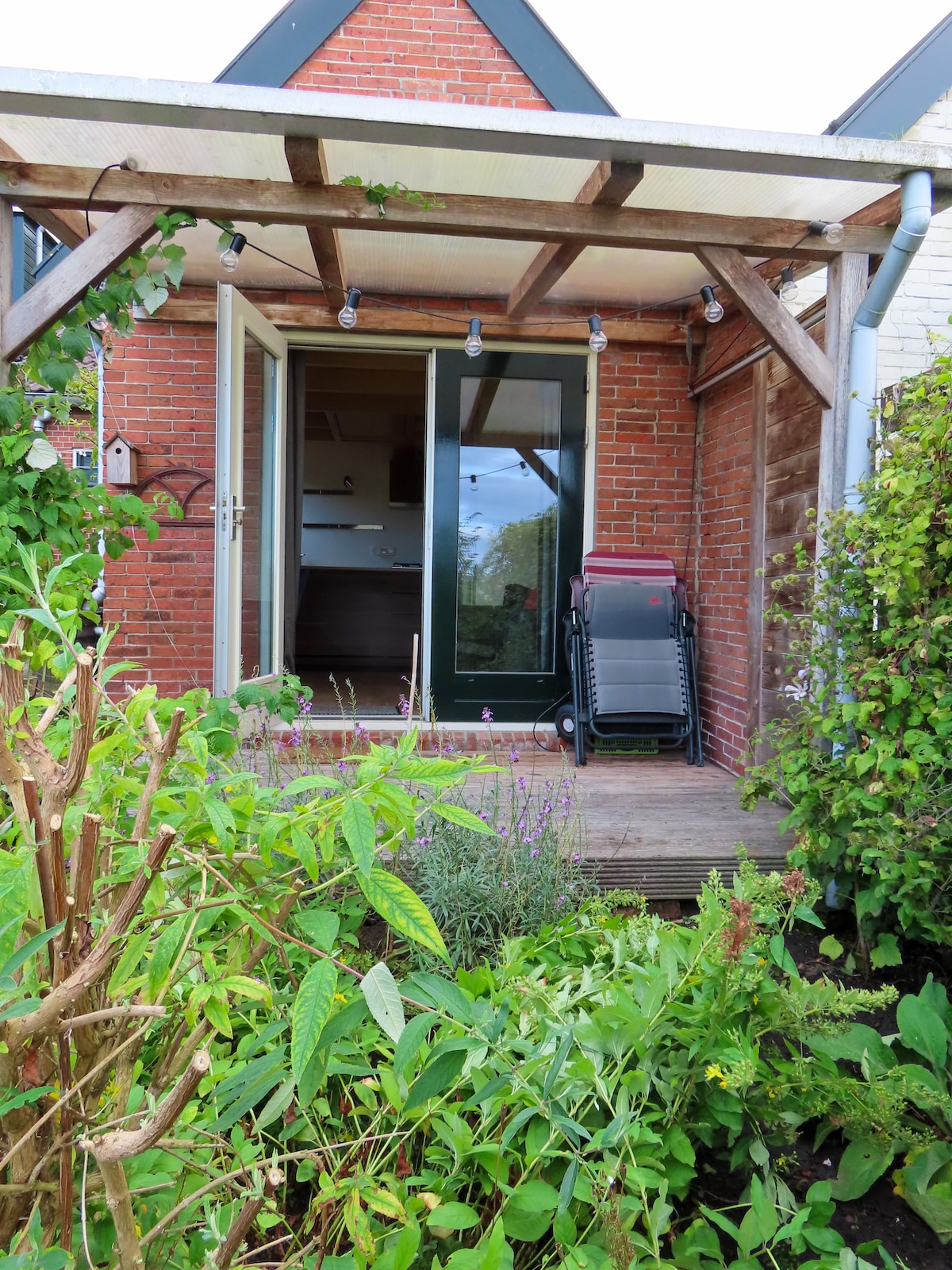 The entrance of the mini-house is framed by vibrant greenery, featuring a wooden pergola with string lights. A sleek, folded stroller is positioned near the door, providing easy access. The interior can be glimpsed through large glass doors, inviting natural light into the space.