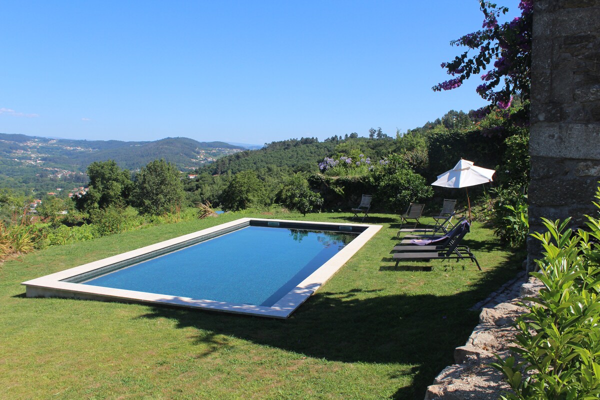 An exclusive swimming pool is framed by a manicured lawn and surrounded by lush greenery, showcasing a serene landscape. A large umbrella offers shade next to two lounge chairs, inviting relaxation. The expansive view of rolling hills is visible in the background under a clear blue sky.